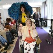 C'est un défilé. Une jeune en Service civique portant une ombrelle bleue pousse le fauteuil roulant d'une dame âgée maquillée et habillé pour l'événement